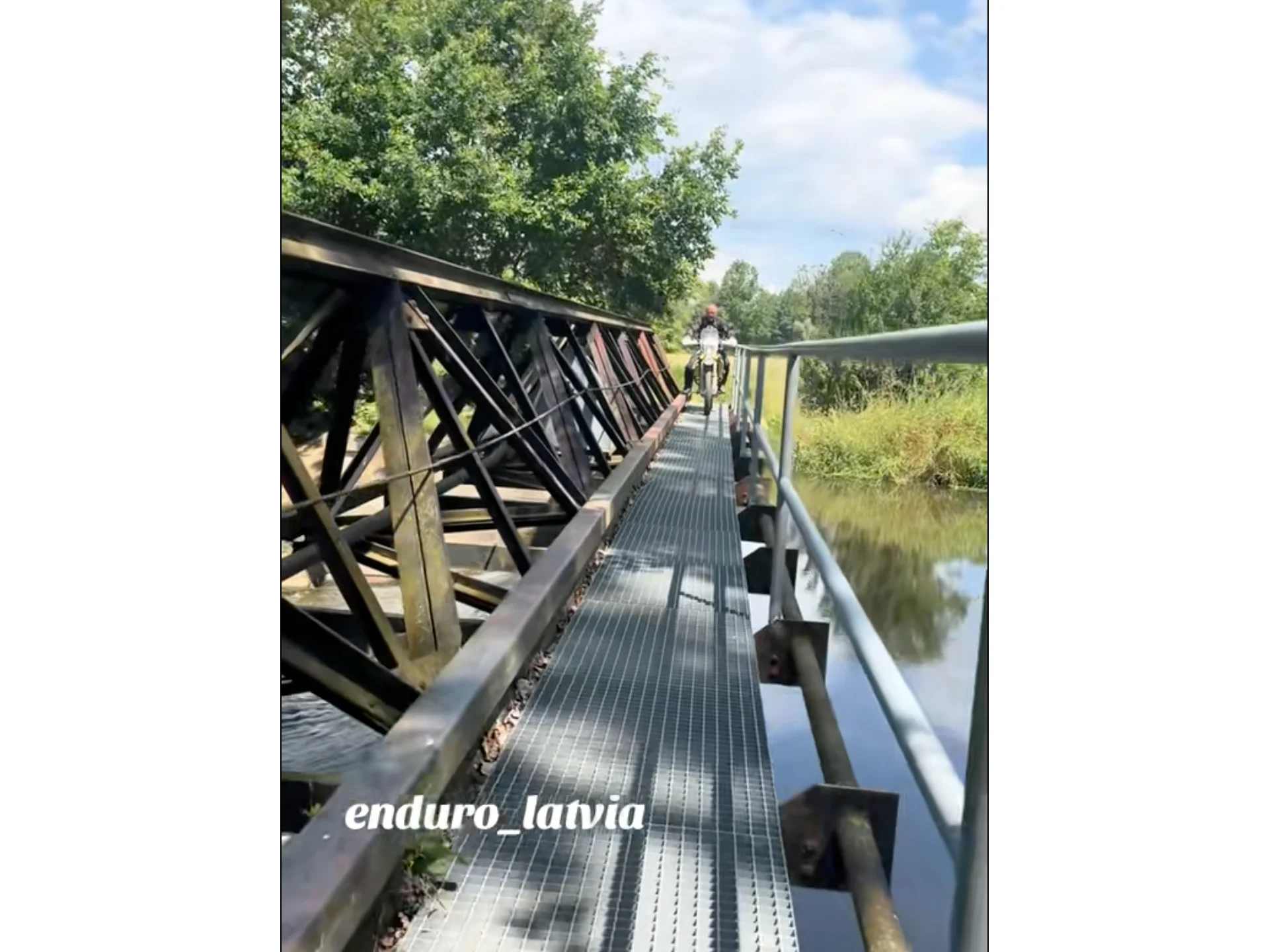 Enduro rider crossing a weathered metal bridge over a tranquil river surrounded by lush green trees
